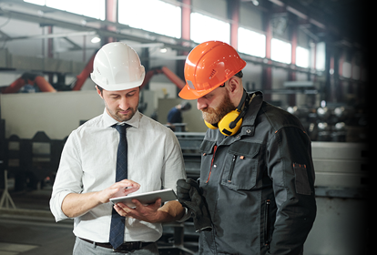 Young master in hardhat and bearded engineer discussing technical sketch on display of tablet in factory workshop