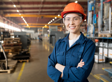 Young smiling female technician in blue uniform, protective eyeglasses and helmet working in modern factory