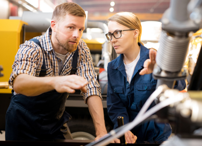 One of young confident technicians pointing at mechanism of industrial equipment during discussion of its qualities with colleague