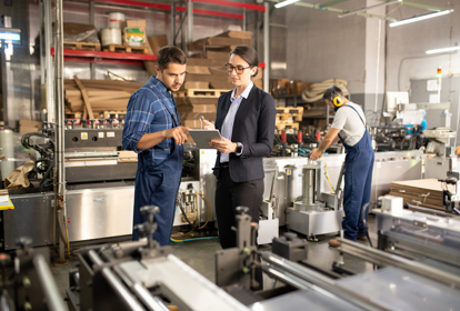 Two young confident workers in a processing factory discussing data on a tablet while standing by one of industrial machines