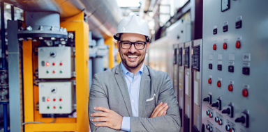 A businessman in grey suit and with helmet on head standing with arms folded next to dashboard inside a Power plant.