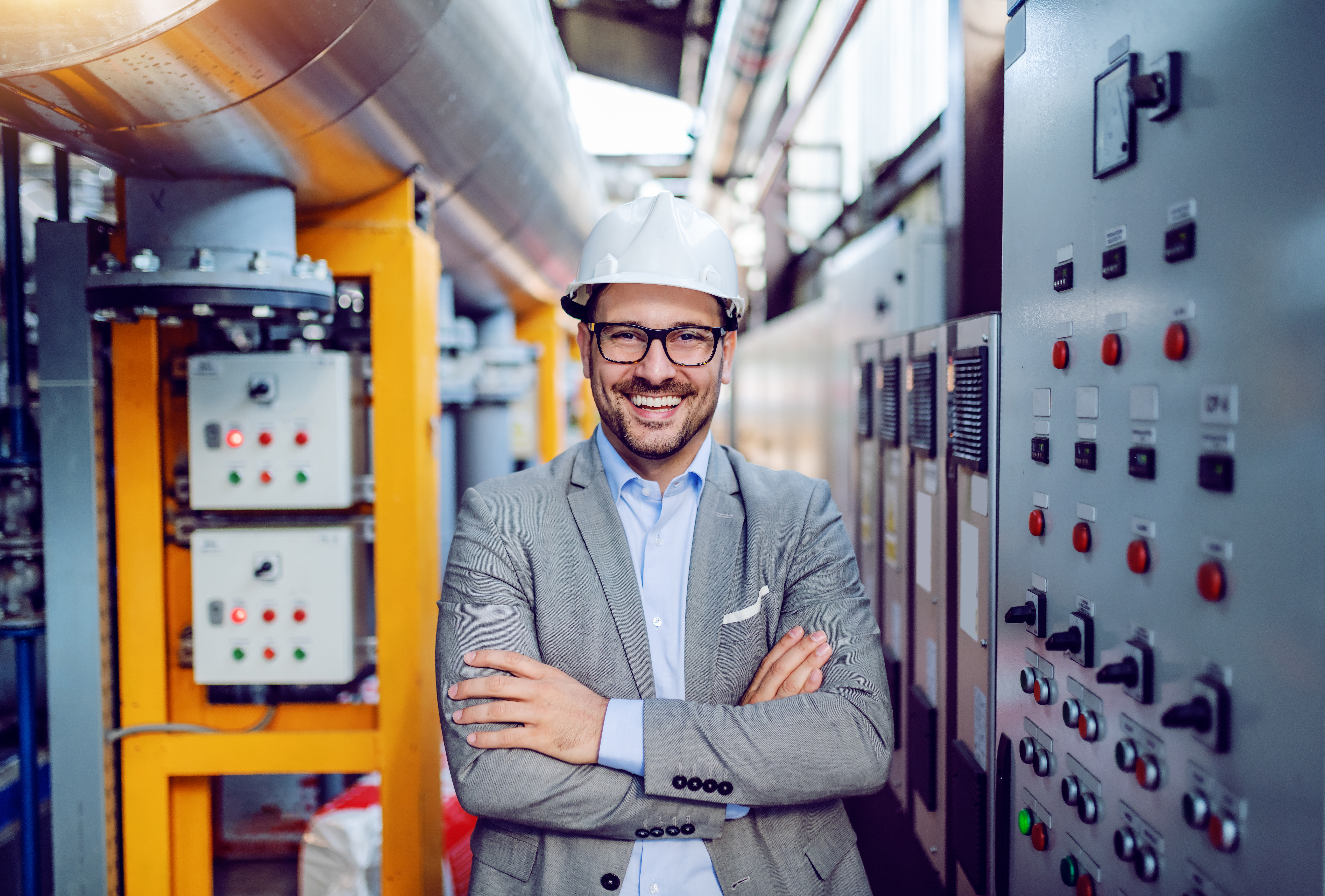 A businessman in grey suit and with helmet on head standing with arms folded next to dashboard inside a Power plant.