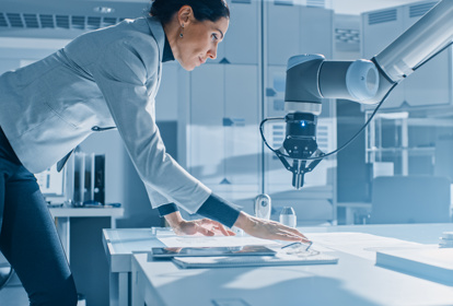 Female Robotics Engineer Leans on the Table Works with Blueprints, Documents and Tablet Computer, She's Programming Robot Arm Movements